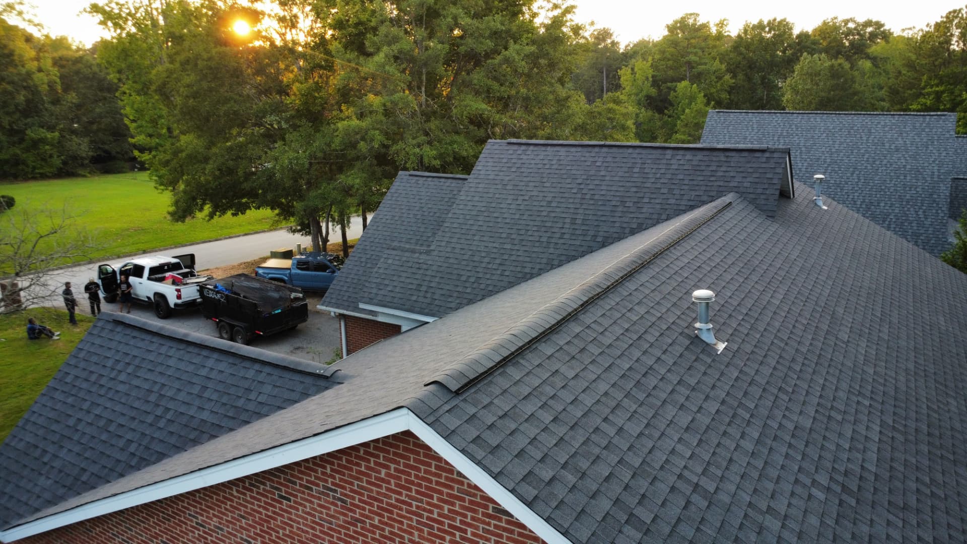 Aerial sunset view of a completed charcoal architectural shingle roof replacement with the Coster Roofing crew and dump trailer on site in Greenwood County SC